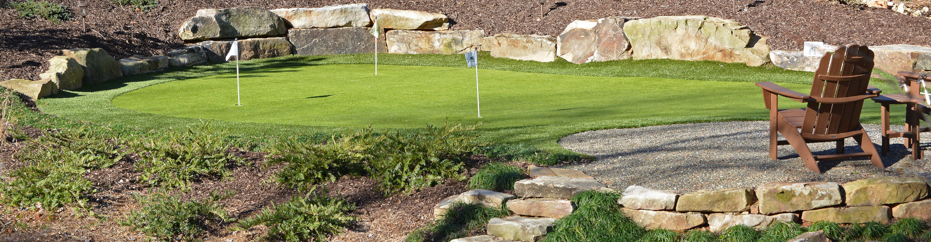 Putting green with three white flag pins framed by large natural boulder retaining walls, stone step pathway, and mulch garden beds